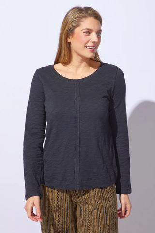 Smiling woman wearing a black long-sleeve scoop neck top with a visible front seam, paired with patterned brown pants, standing against a white background.