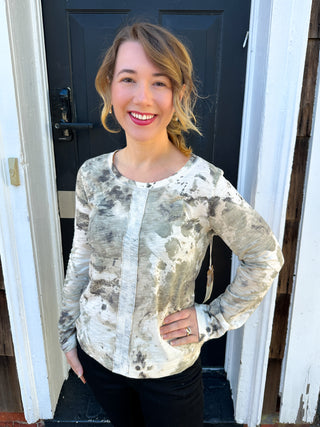 Woman smiling, wearing a women's abstract distressed long sleeve knit top in off-white, grey, and olive green, paired with black pants, standing in front of a dark door.