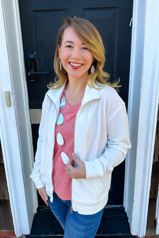 Smiling woman modeling a cream-colored waffle knit full-zip jacket over a peach V-neck shirt and blue jeans, accessorized with a long statement necklace and earrings, standing in front of a dark door.