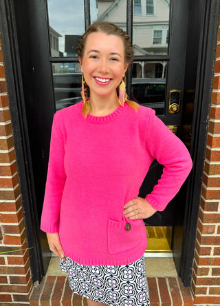 A smiling woman wearing a bright hot pink textured knit long-sleeve crew neck sweater with a decorative side pocket and a wooden button, paired with a black and white floral patterned skort. She stands in front of a dark door.