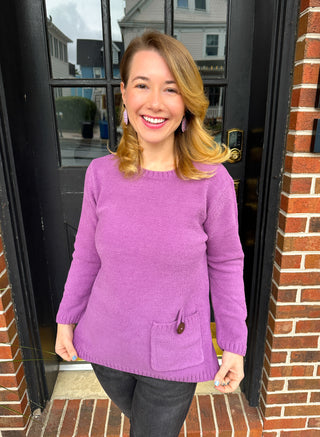 A smiling woman wearing a cozy lavender textured knit crew neck sweater with a decorative brown buttoned pocket, black jeans, and purple earrings, standing in front of a black door and brick wall.