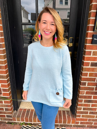 A smiling woman with golden blonde hair models a pastel sky blue chenille sweater, featuring a cozy texture and a unique patch pocket with a prominent brown button, paired with blue jeans against a brick building backdrop.