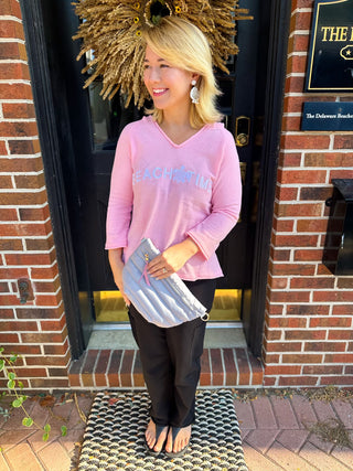 A smiling blonde woman wears a light pink V-neck lightweight knit chenille hoodie with a 'BEACH TIME' and turtle graphic, standing by a dark door with a large golden wreath and holding a grey clutch.