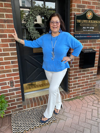 A woman smiling, wearing a bright royal blue chenille knit sweater with 3/4 sleeves and a single buttoned pocket, standing in front of a brick building with a dark door and wreath.