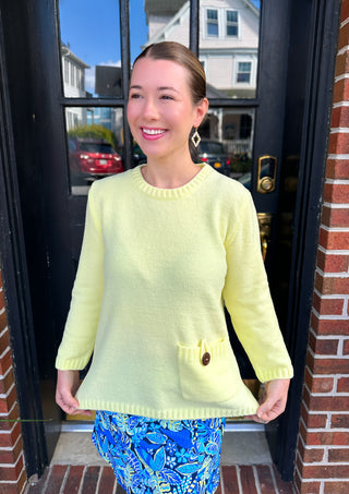 Smiling woman wearing a soft, relaxed-fit lemon yellow crewneck sweater with a decorative brown button, paired with a blue and yellow patterned skort, standing in front of a dark door.