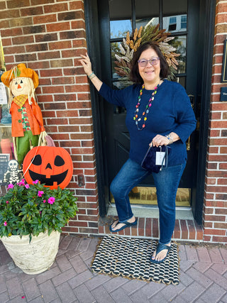 A woman wearing a cozy navy blue soft knit sweater with 3/4 sleeves, a classic crew neck, and a decorative wooden button on a small patch pocket. She is also wearing blue jeans and layered bracelets.