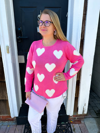 A smiling blonde woman in glasses wears a vibrant hot pink knit sweater adorned with scattered white hearts, paired with white pants, standing in front of a dark wooden door.