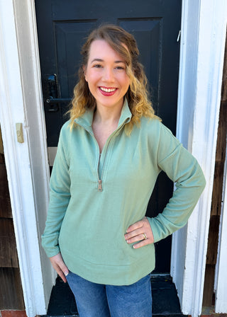 A smiling woman with blonde curly hair wears a light mint green ribbed quarter-zip pullover and blue jeans, standing in front of a black door and white trim.