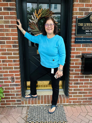 A smiling woman wearing a cozy, vibrant aqua blue textured knit sweater with a front patch pocket and decorative button, standing against a brick wall and dark door with a wreath.